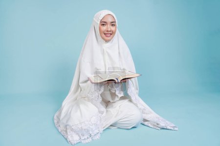 Heureuse jeune femme musulmane asiatique en robe de prière assise et lisant le Coran sur un fond bleu isolé. Concept du Hadj.