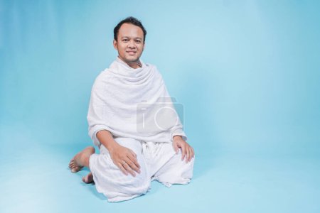 Heureux jeune homme musulman asiatique portant ihram assis sur un fond bleu isolé. Concept du Hadj.