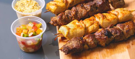 Foto de Brazilian churrasquinho. Kebabs with beef and chicken meat and bacon, known in Brazil by the name of churrasquinho. Wooden board, farofa and vinaigrette in the composition. Above view. - Imagen libre de derechos