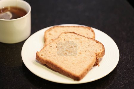 Photo for Two slices of whole grain bread on a small white plate with a cup of tea in the background. Macro photography. Black granite background. - Royalty Free Image
