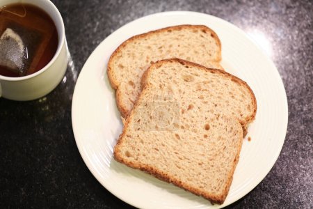 Téléchargez les photos : Deux tranches de pain de grains entiers sur une petite assiette blanche avec une tasse de thé en arrière-plan. Photographie macro. Fond de granit noir. - en image libre de droit