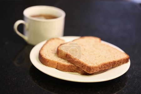 Foto de Dos rebanadas de pan integral en un pequeño plato blanco con una taza de té en el fondo. Fotografía macro. Fondo de granito negro. - Imagen libre de derechos