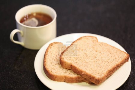 Photo for Two slices of whole grain bread on a small white plate with a cup of tea in the background. Macro photography. Black granite background. - Royalty Free Image