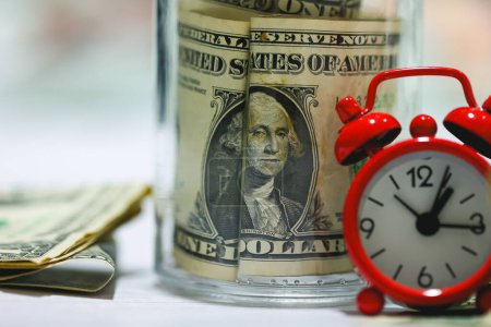 Foto de Billetes de dólar con frasco de vidrio en una mesa de madera con un despertador rojo en la composición. Concepto de ahorro. - Imagen libre de derechos