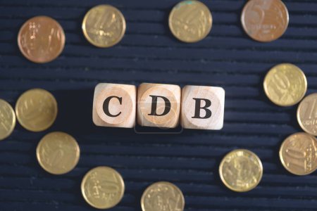 Photo for The acronym CDB for Bank Deposit Certificate in Brazilian Portuguese written on wooden cubes and coins on black background. - Royalty Free Image
