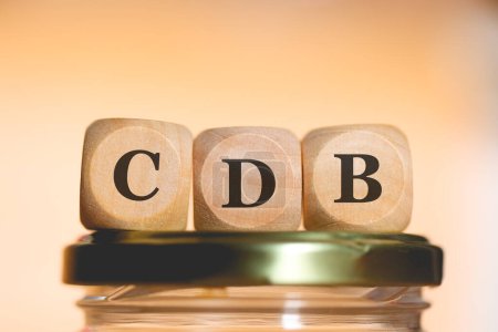 The abbreviation CDB written on wooden dice lying on top of a glass money-saving jar. Studio photo.