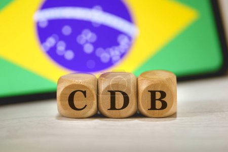 Foto de El acrónimo CDB para Certificado de Depósito Bancario escrito en cubos de madera. Un teléfono celular con la bandera brasileña en la pantalla en la composición. - Imagen libre de derechos