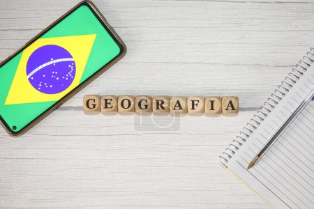Téléchargez les photos : Le texte GEOGRAPHIE en portugais écrit sur des cubes de bois sur une table en bois. Un carnet, un stylo et un téléphone portable avec le drapeau brésilien dans la composition. - en image libre de droit