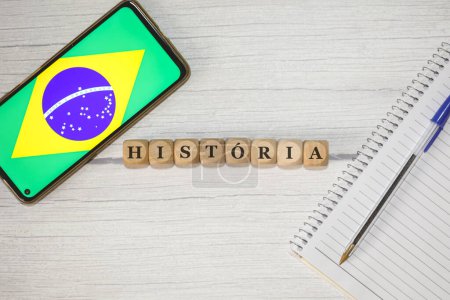 Foto de El texto HISTORIA en portugués escrito sobre cubos de madera sobre una mesa de madera. Un cuaderno, un bolígrafo y un celular con la bandera brasileña en la composición. - Imagen libre de derechos