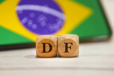 Photo for The acronym DF Federal District in Brazilian Portuguese written on wooden cubes. A cell phone with the Brazilian flag on the screen in the composition. - Royalty Free Image
