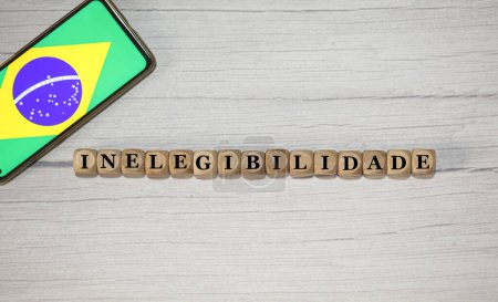 Photo for The text INELIGIBILITY in Brazilian Portuguese written on wooden cubes. A cell phone with the Brazilian flag being shown on the screen in the composition. - Royalty Free Image