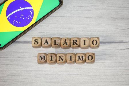 Foto de El texto MÍNIMO Salario en portugués brasileño escrito sobre cubos de madera. Un teléfono celular con la bandera brasileña que se muestra en la pantalla en la composición. - Imagen libre de derechos