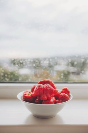 White bowl of organic fresh strawberries on a windowsill with raindrops. Fresh strawberries in the ceramic bawl. Yummy strawberries. 