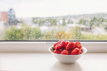 Téléchargez les photos : Bol blanc de fraises fraîches biologiques sur un rebord de fenêtre avec des gouttes de pluie. Fraises fraîches dans le bawl en céramique. Fraises délicieuses. - en image libre de droit