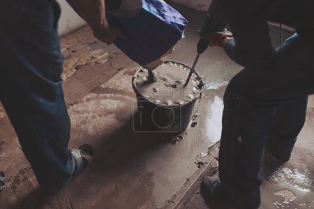 Workers put dry plaster into a bucket. Workers mixing concrete in bucket indoors. A professional craftsman kneads mortar in a bucket with a mixer - adhesive mortar for bricks. 