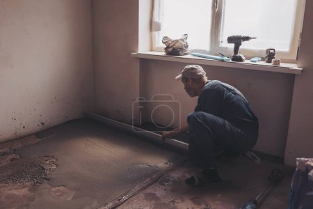 Männlicher Bauarbeiter mit Estrichschiene beim Estrichbodeneinbau im Wohnzimmer. Mann glättet und glättet bei Renovierung Fläche mit gerader Kante in Wohnung.