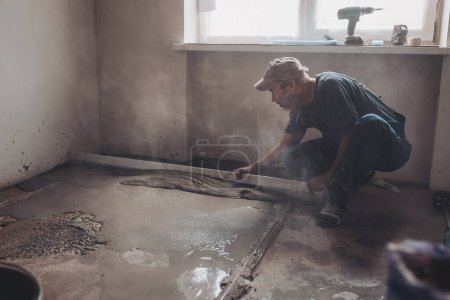 Male construction worker using screed rail while screeding floor in living room. Man flattening and smoothing surface with straight edge in apartment during renovation.