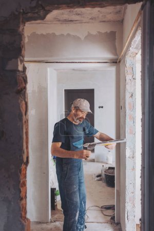 Side view of a plasterer standing in a room in renovation process and using tools for plasterwork and skim coating walls. A worker is renovating house and doing manual building works.