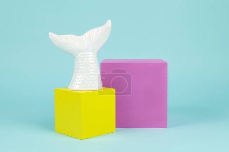Foto de Una cola de sirena en cerámica iridiscente sobre una pila de cubos rosados y amarillos. Fondo turquesa. Fotografía de bodegón mínima y creativa - Imagen libre de derechos
