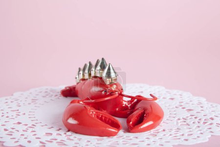 Photo for A punk style lobster wearing riveted spikes like a mohican on a white doily and pink background. Minimal color still life photography. - Royalty Free Image
