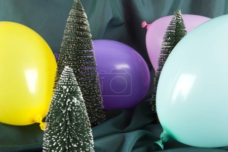 Photo for A composition of snowy Christmas trees surrounded by multicolored balloons on a wavy green background. Minimal color still life photography. - Royalty Free Image