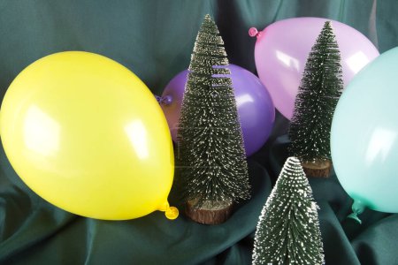 Téléchargez les photos : Une composition d'arbres de Noël enneigés entourés de ballons multicolores sur un fond vert ondulé. Photographie minime couleur nature morte. - en image libre de droit