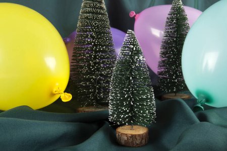 Téléchargez les photos : Une composition d'arbres de Noël enneigés entourés de ballons multicolores sur un fond vert ondulé. Photographie minime couleur nature morte. - en image libre de droit