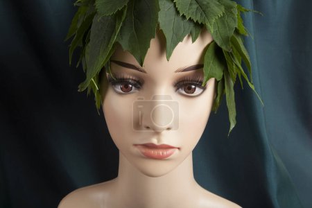 Téléchargez les photos : Un mannequin de vitrine coiffé de feuilles et représentant une déesse de la nature devant un rideau vert. Photographie minime couleur nature morte. - en image libre de droit
