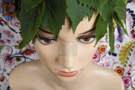 Photo for A display mannequin wearing hair of leaves and representing a nature goddess in front of a floral pattern curtain. Minimal color still life photography. - Royalty Free Image