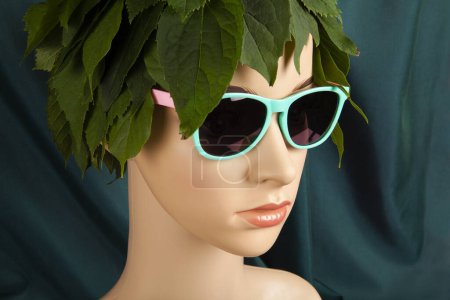 Photo for A display mannequin wearing hair of leaves and sunglasses, representing a nature goddess in front of a green curtain. Minimal color still life photography. - Royalty Free Image