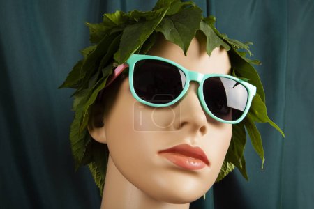 Téléchargez les photos : Un mannequin de vitrine coiffé de feuilles et de lunettes de soleil, représentant une déesse de la nature devant un rideau vert. Photographie minime couleur nature morte. - en image libre de droit
