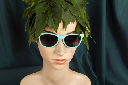 Foto de Un maniquí de exhibición con pelo de hojas y gafas de sol, que representa a una diosa de la naturaleza frente a una cortina verde. Fotografía de bodegón de color mínimo. - Imagen libre de derechos