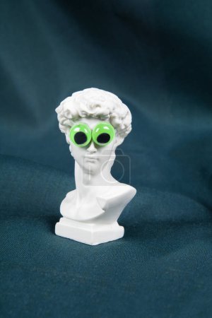 Photo for A bust of David towering on a wavy green curtain with fake bulging eyes. Comic scenography. Minimal color still life photography - Royalty Free Image