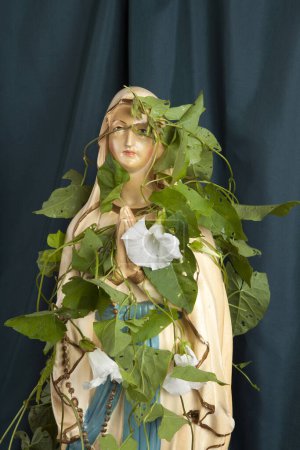 Téléchargez les photos : Une statuette de la Vierge Marie couverte d'asclépiade devant un rideau vert ondulé. une métaphore du temps et de sa poésie. Photographie minime de nature morte - en image libre de droit