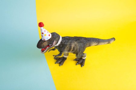 Foto de Un dinosaurio, vestido con encaje y un sombrero de fiesta de papel, en una fiesta de cumpleaños sobre un fondo amarillo y azul. Mínimo creativo bodegón fotografía colorida - Imagen libre de derechos
