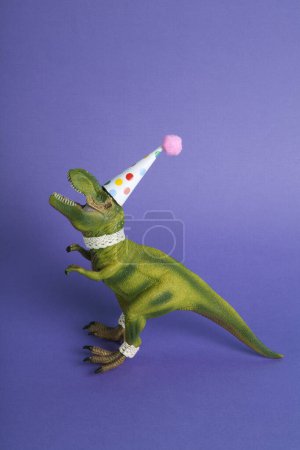 Téléchargez les photos : Un dinosaure, habillé en dentelle et un chapeau de fête en papier, lors d'une fête d'anniversaire sur un fond violet. Minimal créatif nature morte photographie colorée - en image libre de droit