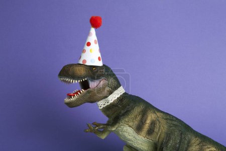 Téléchargez les photos : Un dinosaure, habillé en dentelle et un chapeau de fête en papier, lors d'une fête d'anniversaire sur un fond violet. Minimal créatif nature morte photographie colorée - en image libre de droit