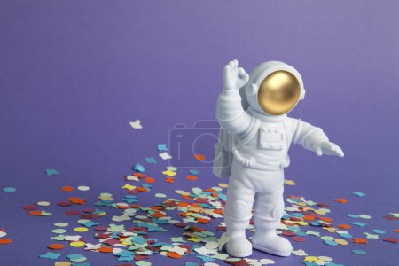 Téléchargez les photos : Une figurine astronaute dans une combinaison spatiale explorant un fond violet uni sur un tapis de confettis. Minimal créatif nature morte photographie colorée - en image libre de droit