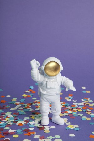 Téléchargez les photos : Une figurine astronaute dans une combinaison spatiale explorant un fond violet uni sur un tapis de confettis. Minimal créatif nature morte photographie colorée - en image libre de droit