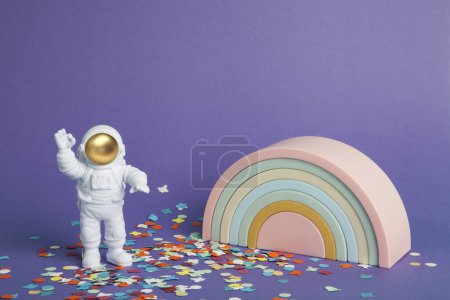 Téléchargez les photos : Une figurine d'astronaute dans une combinaison spatiale explorant pour le trésor un fond violet uni sur un tapis de confettis et devant un arc-en-ciel en plastique. Minimal créatif nature morte photographie colorée - en image libre de droit
