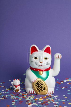 Foto de Una pareja madre y el bebé de Maneki-neko gato de plástico, Simbolizando la suerte y la riqueza, en una alfombra de confeti y un fondo violeta pop. Mínimo color de moda bodegón fotografía - Imagen libre de derechos