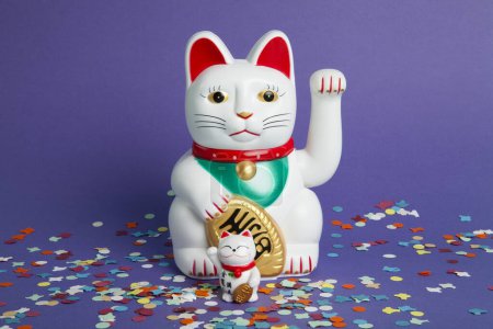 Foto de Una pareja madre y el bebé de Maneki-neko gato de plástico, Simbolizando la suerte y la riqueza, en una alfombra de confeti y un fondo violeta pop. Mínimo color de moda bodegón fotografía - Imagen libre de derechos