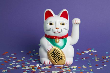 Téléchargez les photos : Un chat en plastique Maneki-neko blanc, symbolisant chance et richesse, sur un tapis de confettis et un fond violet pop. Minimale couleur tendance photographie nature morte - en image libre de droit