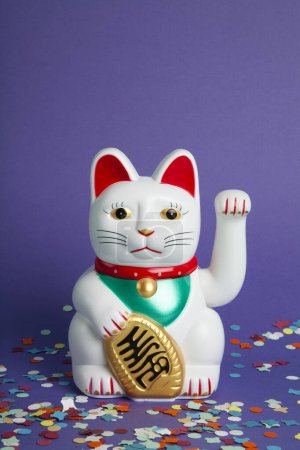 Téléchargez les photos : Un chat en plastique Maneki-neko blanc, symbolisant chance et richesse, sur un tapis de confettis et un fond violet pop. Minimale couleur tendance photographie nature morte - en image libre de droit