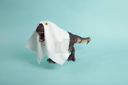 Photo for A plastic t-rex dinosaur figurine disguised as a ghost with a sheet and false eyes on a turquoise background. Minimalist, trendy still life photography. - Royalty Free Image