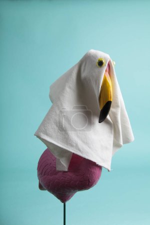 Photo for A plastic flamingo decoration disguised as a ghost with a sheet and false eyes on a turquoise background. Minimalist, trendy still life photography. - Royalty Free Image