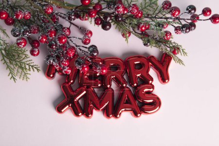 Foto de Un adorno de Navidad de plástico rojo brillante con un texto que dice Feliz Navidad. Una rama de acebo con sus bayas rojas enmarcando la parte superior de la foto. Minimalista, fotografía bodegón de moda. - Imagen libre de derechos