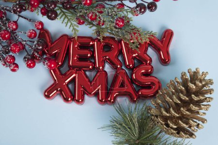 Téléchargez les photos : Un ornement de Noël en plastique rouge brillant avec un texte disant joyeux Noël. Une branche de houx avec ses baies rouges encadrant le haut de la photo et un cône de pin doré. Fond bleu. Photographie de nature morte minimaliste et tendance. - en image libre de droit