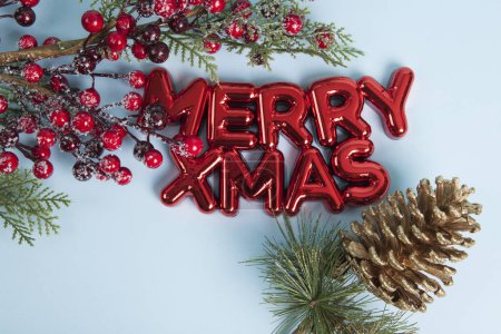 Photo for A glossy red plastic Christmas ornament featuring a text saying merry xmas. A branch of holly with its red berries framing the top of the photo and a golden pine cone. Blue background. Minimalist, trendy still life photography. - Royalty Free Image