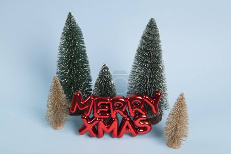 Foto de Un adorno de Navidad de plástico rojo brillante con un texto que dice feliz Navidad frente a un bosque de árboles de navidad verdes y dorados. Minimalista, fotografía bodegón de moda. - Imagen libre de derechos